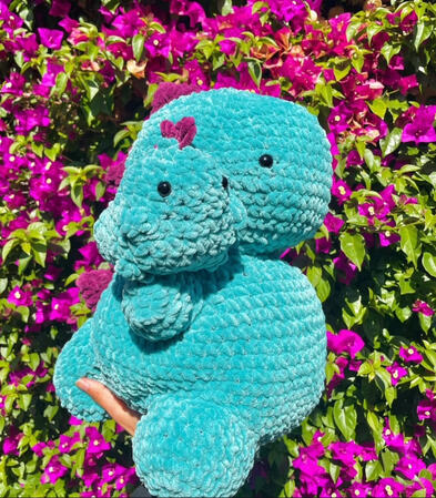 a large crocheted teal dinosaur with purple spikes holding a small crocheted teal dinosaur