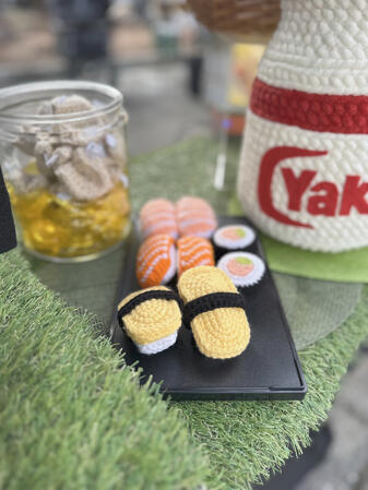 a plate of crocheted sushi keychains. tamago nigiri, shrimp nigiri, salmon nigiri, salmon and avocado maki. crocheted fortune cookies and a yakult bottle are in the background.